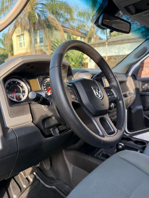 Interior view of a car dashboard with steering wheel, gauges, and windshield view of residential area with trees and houses