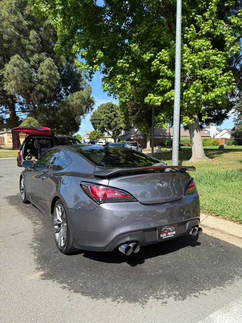 Gray sports coupe parked on residential street with open trunk, surrounded by trees and houses